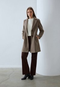 Femme se tient contre un mur blanc, portant un manteau en laine taupe, un pull crème, un pantalon marron foncé et des bottes à talons noires avec les mains dans les poches du manteau.