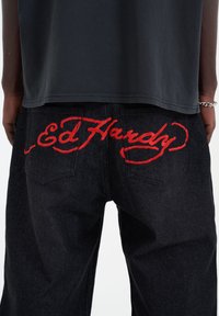 Černé džíny z denimu s červeným vyšívaným textem "Ed Hardy" na zadní straně. Klasický design s pěti kapsami a mírně volným střihem.