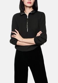 Haut noir zippé avec col, présentant des manches froncées et un détail textuel sur la fermeture éclair. Porté avec un pantalon noir à rayures verticales.