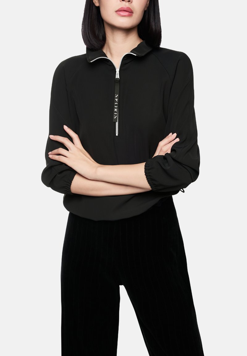 Haut noir zippé avec col, présentant des manches froncées et un détail textuel sur la fermeture éclair. Porté avec un pantalon noir à rayures verticales.