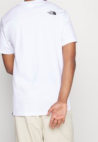 Camiseta de algodón blanca con un logo negro en la parte superior de la espalda, de manga corta y un ajuste relajado, combinada con pantalones de color claro.