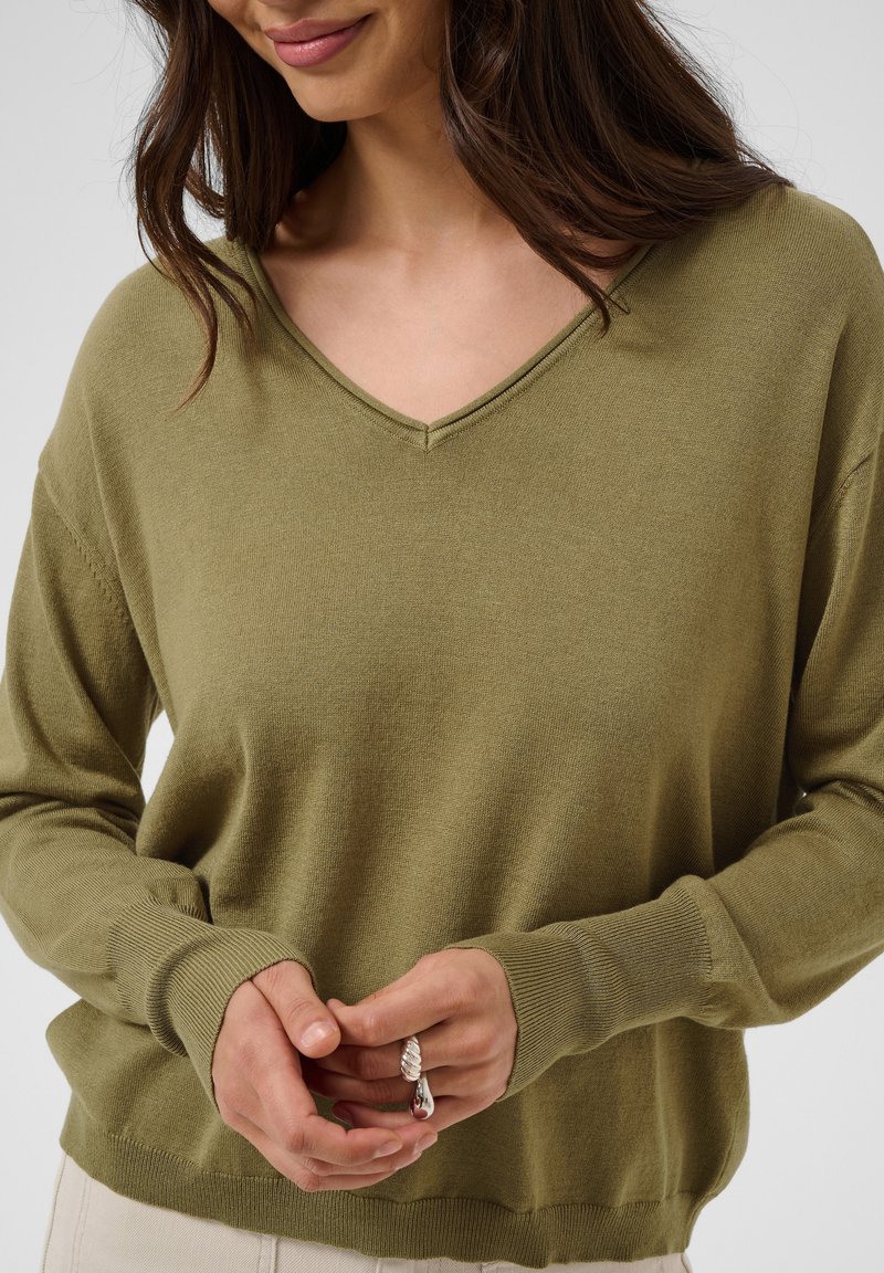 Femme portant un pull à col en V vert olive, montrant ses mains jointes devant elle tenant une bague en argent avec un pendentif.
