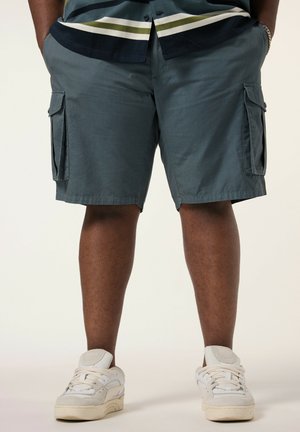 Persoon draagt donkergrijze cargoshorts, witte sneakers en een gestreept shirt met handen in de zakken, tegen een effen achtergrond.