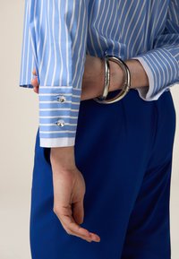 Camicia a righe blu chiaro con polsini bianchi, caratterizzata da due bottoni di cristallo sul polsino; una mano tiene dei braccialetti d'argento. Pantaloni blu sullo sfondo.