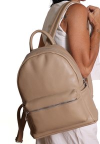 Beige leren rugzak met een afgeronde vorm, dubbele schouderbanden, handvat bovenop en een voorste ritszak. Getextureerd oppervlak met glanzende accenten.