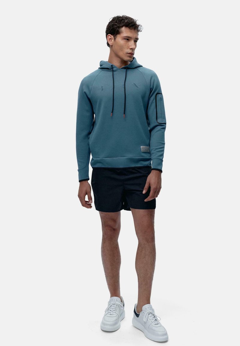 Kapuca majica v teal barvi s nastavljivimi vrvicami, teksturiranimi detajli in žepom na zadrgo na rokavu. V kombinaciji z črnimi kratkimi hlačami in belimi športnimi copati.