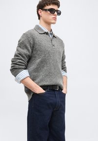 Άνδρας που φοράει μαύρα γωνιώδη γυαλιά ηλίου, γκρι πουλόβερ πάνω από μπλε ριγέ πουκάμισο, σκούρο μπλε τζιν, με τα χέρια στις τσέπες, μπροστά σε απλό φόντο.