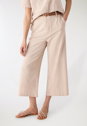 Pantaloni beige a gamba larga e taglio corto abbinati a una cintura marrone e sandali piatti beige con cinturini indossati da una persona in piedi.