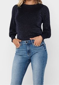 Frau trägt einen dunkelblauen Langarmpullover und hellblaue, hoch sitzende Jeans mit den Händen in den vorderen Taschen.