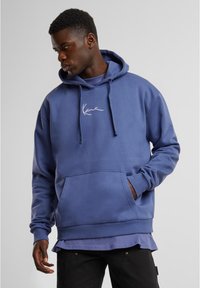 Felpa blu in cotone con tasca frontale, cappuccio con lacci e logo in corsivo bianco. Presenta un orlo e polsini a costine, indossata sopra una maglietta più chiara.