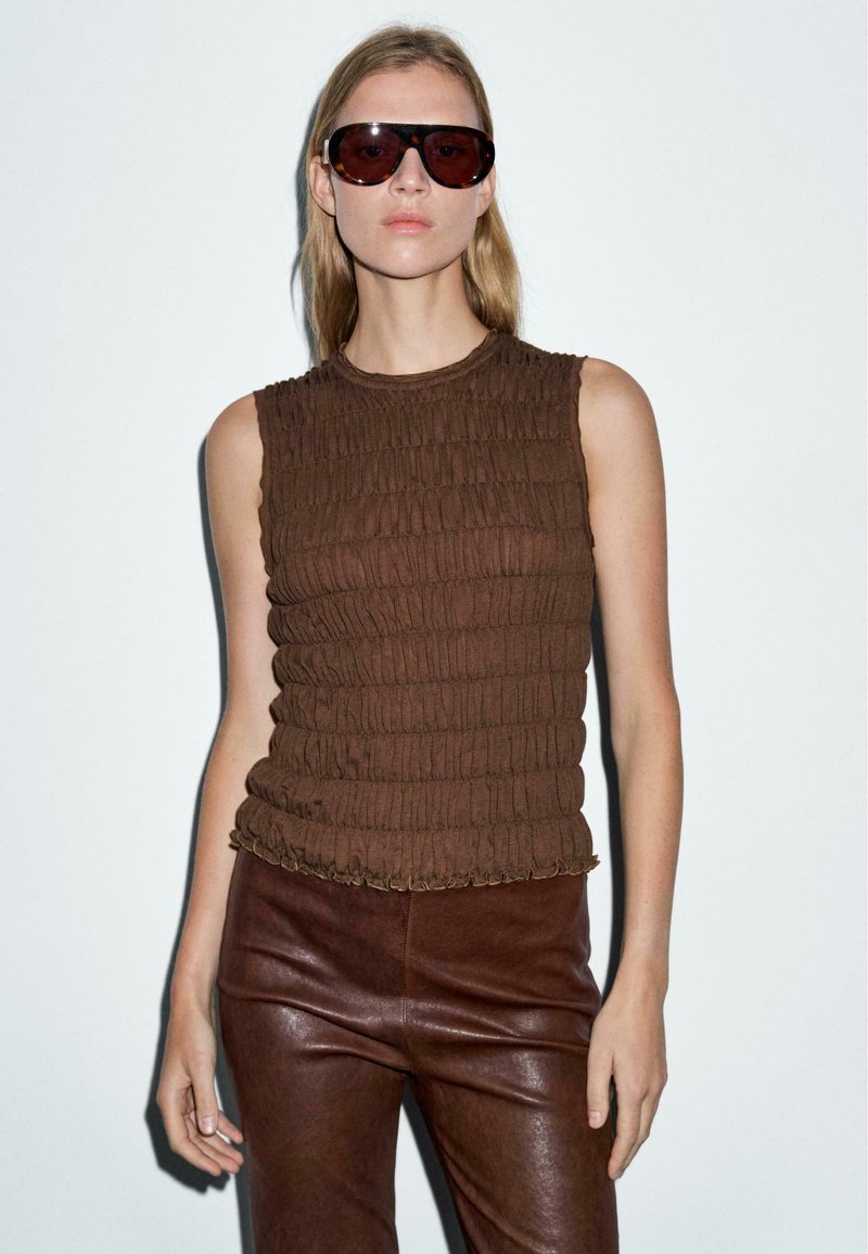 Top senza maniche di colore marrone con dettagli plissettati, abbinato a pantaloni in pelle marrone e occhiali da sole oversize di colore scuro. Design minimalista.