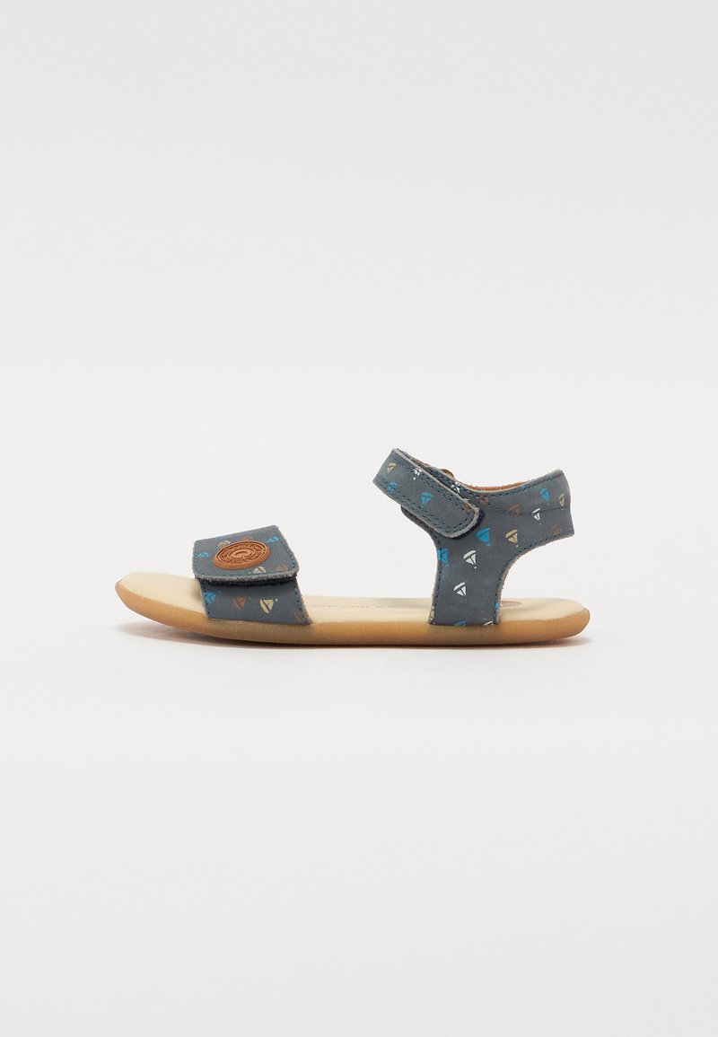 Blå sandaler med et geometrisk mønster og klemmestropper, med et rundt logo foran og en flat lysebrun såle.