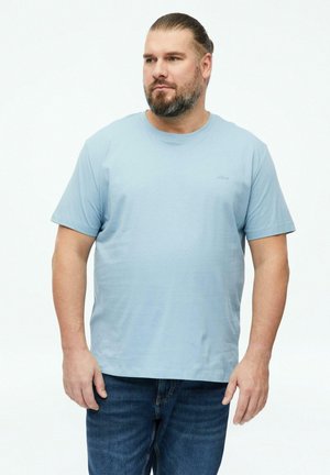 Mann mit Bart und zurückgebundenem Haar, der ein hellblaues kurzärmeliges T-Shirt und dunkelblaue Jeans trägt, steht vor einem einfarbigen weißen Hintergrund.