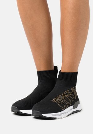 Sorte sokkestil sneakers med hvide såler, ribstrikkede ankelfløjter og gyldent "Versace Jeans Couture" logo på ydersiden, båret på bare ben.