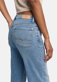 Ljust blå denimjeans med hög midja, som har en läderetikett på baksidan och dekorativ broderi i ett vågigt mönster på fickan.