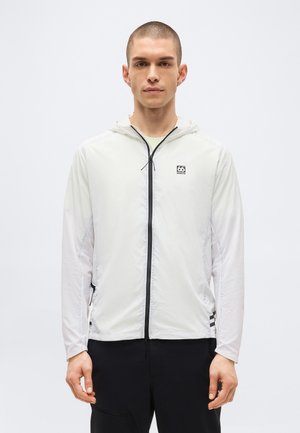 Jeune homme portant une veste à capuche blanche zippée avec une fermeture éclair noire et un logo sur le côté gauche de la poitrine, debout devant un fond clair uni.