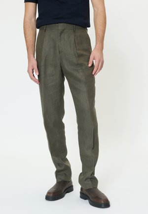 Mand iført olivengrønne plisserede bukser og mørkebrune læderstøvler, stående mod en ensartet lys baggrund.