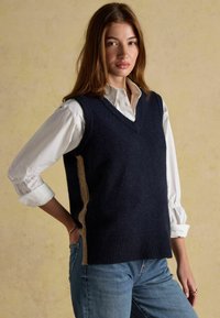 Granatowy bezrękawnik z dekoltem w kształcie V i prążkowanym wykończeniem, z kontrastującymi jasnobrązowymi bocznymi panelami. Noszony na białej koszuli z kołnierzykiem.