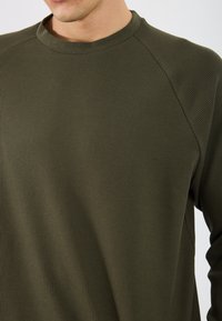 Olīvu zaļš ribots liela garuma krekls ar apaļu kakla izgriezumu un raglāna piedurknēm. Auduma tekstūra ir nedaudz izcilāta, sniedzot unikālu izskatu.