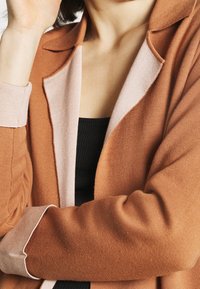 Cardigan de color naranja óxido con una textura suave y lisa. Detalles en rosa claro en el cuello y los puños. Parte superior negra con relieve visible debajo.