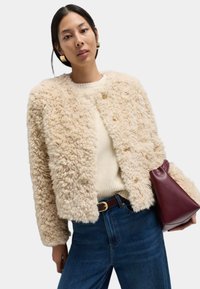Flauschige beige Jacke mit rundem Ausschnitt und goldenen Knöpfen, kombiniert mit einem cremefarbenen Pullover und blauen Jeans. Hält eine kastanienbraune dreieckige Tasche.