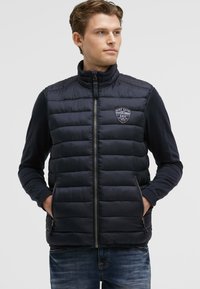 Gilet imbottito blu con zip frontale, colletto alto e patch con logo. Presenta un design trapuntato, tasche laterali e una texture liscia e lucida.