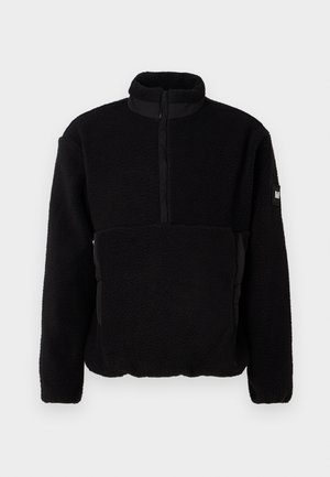 Svart fleece-pullover med høy krage, halv glidelås foran og glatte svarte detaljer. Har en strukturert overflate og logo-lapp på ermene.