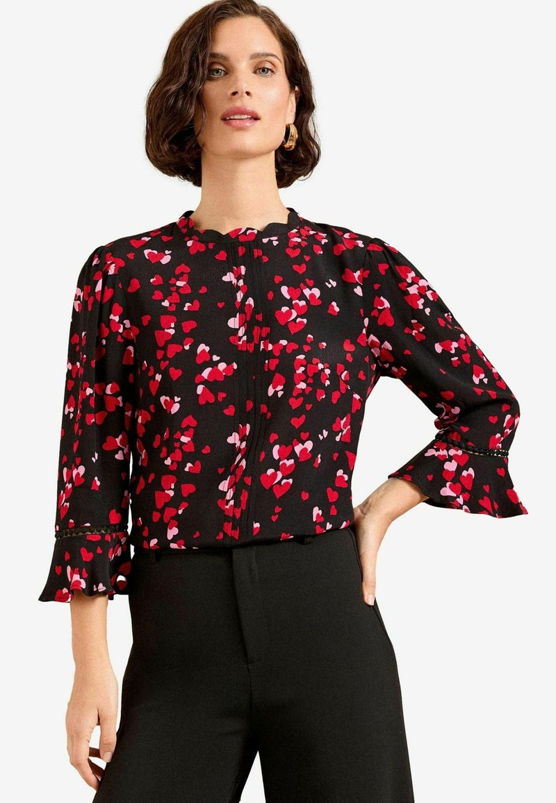 Schwarze Bluse mit roten und rosa Herzmustern, mit langen Ärmeln und Rüschenausschnitten sowie einem runden Ausschnitt, Knopfverschluss vorne.