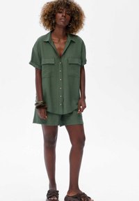 Vrouw met krullend haar, gekleed in een olijfgroene blouse met knopen en bijpassende short, geaccessorieerd met armbanden en bruine slide slippers.