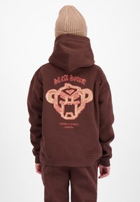 Persoon draagt een bruine hoodie met een abstracte apengezicht en de tekst "BLCK BNNS" en "VIVA LA VIDA" op de achterkant, gecombineerd met bijpassende broek en een beige gebreide muts.