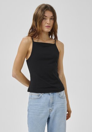 Femeie cu păr lung și brunet, purtând un top negru fără mâneci și blugi bleu deschis cu talie înaltă, pe un fundal gri simplu.