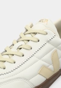Baltas ādas sporta kurpes ar bēšiem rāvējslēdzējiem un sānu logotipu, ar brūnu gumijas zoli un zigzaga šuvēm uz augšdaļas.