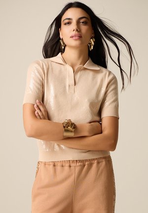 Donna con lunghi capelli scuri che indossa una maglia beige a maniche corte con paillettes, pantaloni beige, orecchini dorati vistosi e un ampio braccialetto d'oro, con le braccia incrociate.