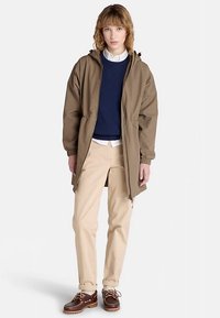 Brązowa kurtka z kapturem i elastycznymi mankietami, noszona na granatowym swetrze i białej koszuli. Dopełnieniem są jasnobeżowe spodnie i ciemnobrązowe buty żeglarskie.