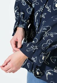 Camicia navy con motivo floreale bianco, dotata di polsini elasticizzati e vestibilità comoda. Tessuto in cotone con texture liscia. Dettaglio del colletto a punta.