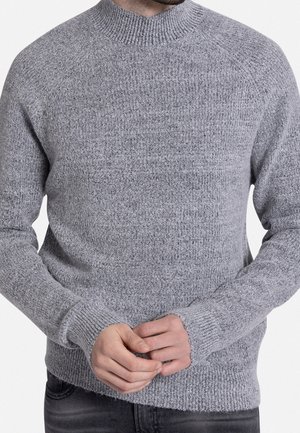 Mand iført en lysegrå, tekstureret striktrøje med lange ærmer, stående med hænderne foldet foran, delvis udsigt fra nakken til hoften.