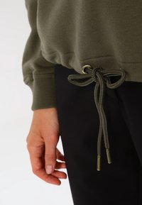 Felpa verde oliva con orlo regolabile, caratterizzata da occhielli in metallo e lacci intrecciati. Il tessuto è morbido e texturizzato, abbinato a pantaloni neri.