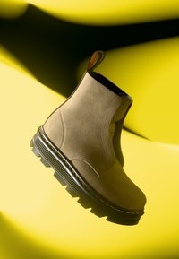 Stivale alla caviglia in suede beige con suola nera spessa e cuciture gialle, mostrato su uno sfondo giallo brillante con ombre.