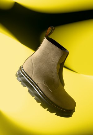 Botín de ante marrón claro con suela negra gruesa y costuras amarillas, presentado sobre un fondo amarillo brillante con sombras.