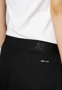 Persona che indossa pantaloni atletici neri perforati con la scritta "DRI-FIT" e una cintura nera che mostra in modo sfocato lo slogan "JUST DO IT", abbinati a una maglietta bianca.