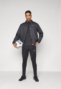 Sort vindjakke med logo, sort crew neck t-shirt, sorte sportbukser med logo, holder en hvid fodbold med blå accenter.