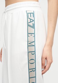 Λευκά παντελόνια με κορδόνι και κάθετη λωρίδα με το λογότυπο EA7 Emporio Armani στο πλάι, φορεμένα από άτομο με ορατό χέρι και κάτω μέρος μέσης.