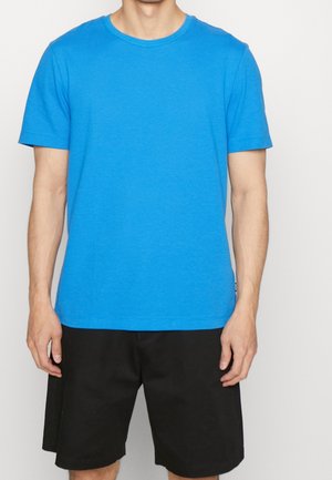 Mężczyzna w jaskrawo niebieskim teksturowanym t-shircie z krótkim rękawem oraz luźnych czarnych szortach do kolan, stojący na gładkim jasnym tle.