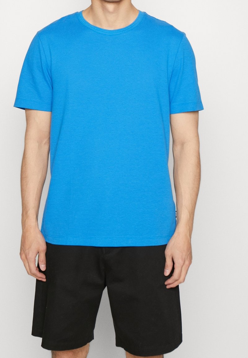 Homme portant un t-shirt bleu vif texturé à manches courtes et un short noir ample jusqu'aux genoux, debout devant un fond clair uni.