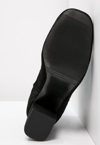 Chaussure en daim noir avec une semelle en caoutchouc texturée et plate. Le design présente un capuchon de talon lisse et une silhouette épurée.