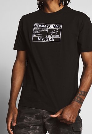 Homme portant un t-shirt noir Tommy Jeans avec des textes graphiques blancs et des symboles, associé à un pantalon noir à motifs, montrant un tatouage sur le bras.