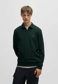 Donkergroene pullover met een hoge kraag, halve rits en een logo op de borst. Gemaakt van een gladde stof, met een getailleerd ontwerp en lange mouwen.