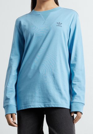 Personne portant un haut à manches longues Adidas bleu clair avec un logo sur la poitrine et un pantalon foncé sur un fond clair uni.