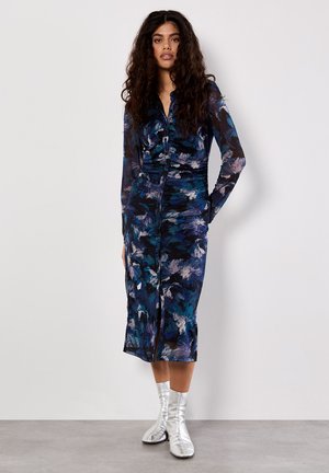 Robe à manches longues avec un motif floral dans des tons bleu marine, bleu et violet. Présente un design à boutons sur le devant et une fente, associée à des bottes argentées.