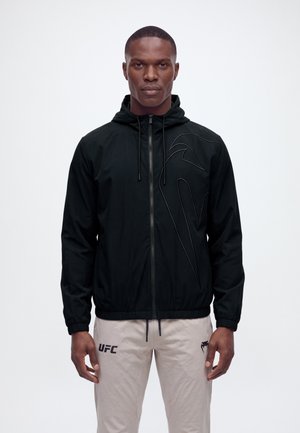Un modelo masculino está de pie mirando al frente, con una sudadera negra con capucha y cremallera y unos pantalones claros con el logotipo de UFC, sobre un fondo blanco liso.